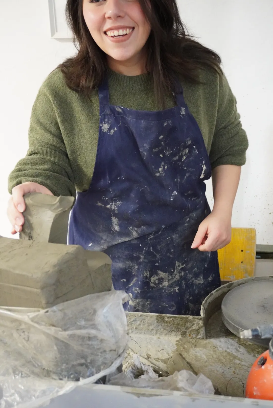 Frida sonríe en el taller sosteniendo una placa de barro, con el mandil manchado